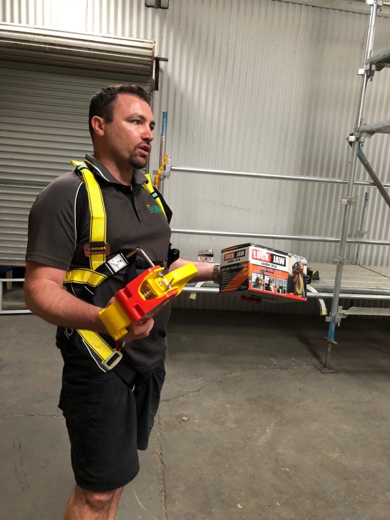 A man in a safety harness holds a Lock Jaw Ladder Grip and its packaging in a warehouse setting.