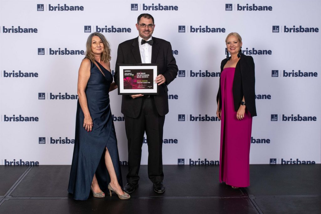 Gail Bray and Craig Charlton receiving an award, with a woman in pink, at a "Brisbane" branded event.