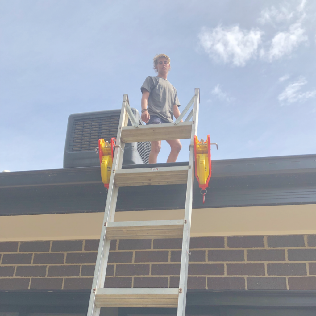 A person stands on a ladder secured with Lock Jaw Ladder Grips, with a brick building and blue sky in the background.