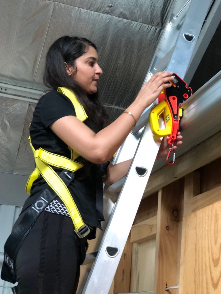 A person in a safety harness holds a Lock Jaw Ladder Grip and its packaging in a warehouse setting.