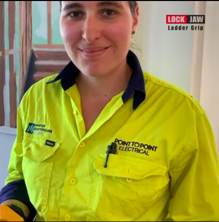 A woman with a bright smile, wearing a black 'Point to Point Electrical' shirt, holds a ladder with a Lock Jaw Ladder Grip.