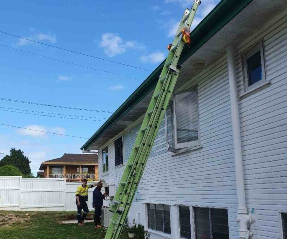 Two workers positioning an extension ladder against a house with white siding and green gutters.