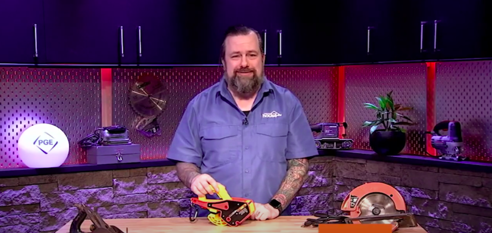 A light-skinned man with a beard and tattoos holds a Lock Jaw Ladder Grip in a studio setting with various tools.